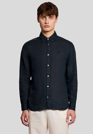 Junger Mann, der ein enganliegendes schwarzes Hemd mit Kragen und cremeweiße Hose trägt, steht vor einem grauen Hintergrund.