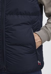 Mørkeblå puffervest med glidelåslommer og myk tekstur. Har en grå langermet skjorte under, som viser ribbede mansjetter. Rød og hvit glidelåspull.