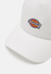 Gorra de algodón blanca con visera curva. Presenta el logotipo bordado de "Dickies" en negro, rojo y amarillo. Tejido texturizado con detalles de costura visibles.