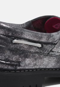 Grauer Textilschuh mit metallischem Finish, kontrastierenden weißen Schnürsenkeln und schwarzer Gummisohle mit sichtbaren Nähten und Ösen-Details.