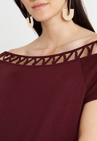 Burgundy topp med avskuren axeldesign, som har ett utskärningsmönster längs halsringningen. Slät tyg med korta ärmar.