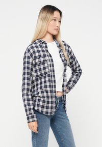 Jauna sieviete ar gariem blondiem matiem, valkājot tumši zilu rūtainu kreklu virs baltas īsās blūzes un ziliem džinsiem, stāv ar vienu roku uz gurna.