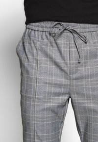 Person wearing gray plaid trousers with a drawstring waist and pockets, paired with a black top.