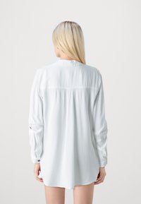 Weißes langärmliges Shirt mit lockerem Schnitt, das einen Schulterpasse-Naht, Knopfärmelakzente und glattes, leichtes Material aufweist.