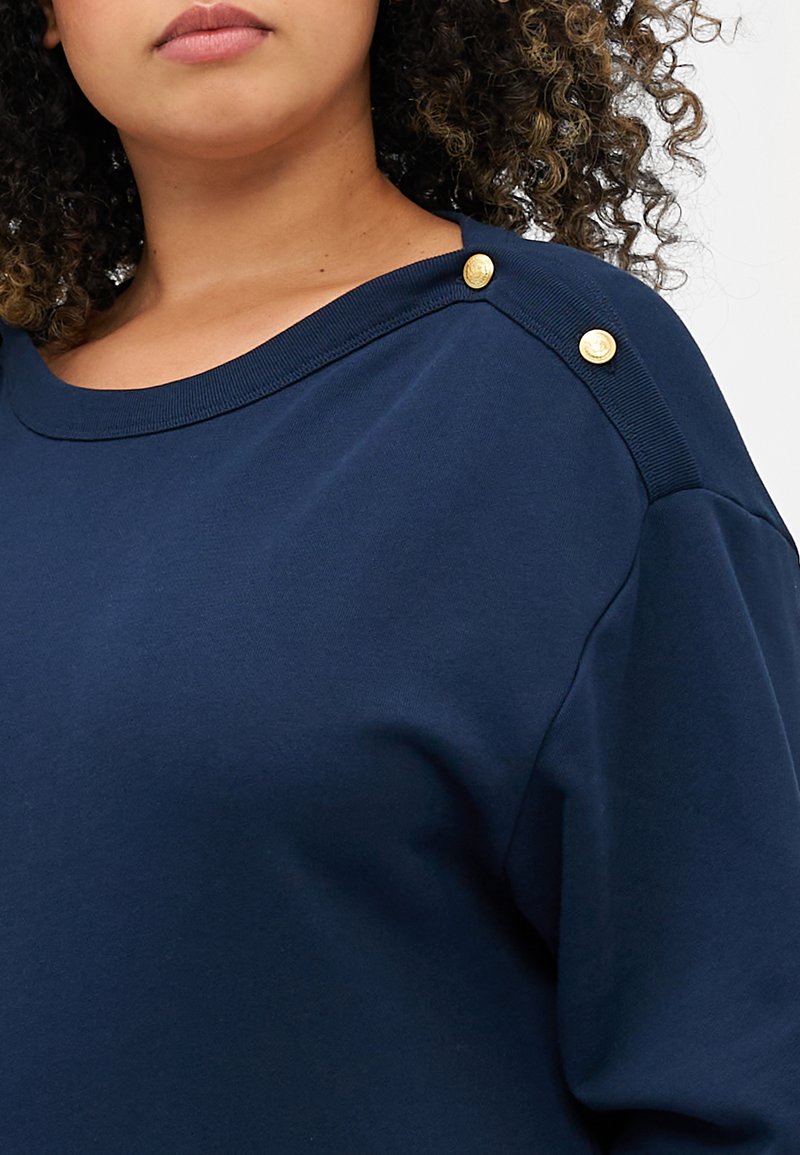 Navyblauwe sweater gemaakt van zachte stof, met een brede halslijn en decoratieve gouden knopen op de linker schouder. Glad textuur.