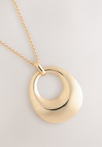 Goldene Halskette mit einem polierten, asymmetrischen runden Anhänger mit großem zentralem Loch, auf hellem Hintergrund liegend.