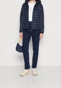 Donna che indossa un giubbotto imbottito blu navy, top bianco, jeans blu scuro, stivali con plateau color crema, e tiene in mano una piccola borsa blu navy con tracolla gialla.