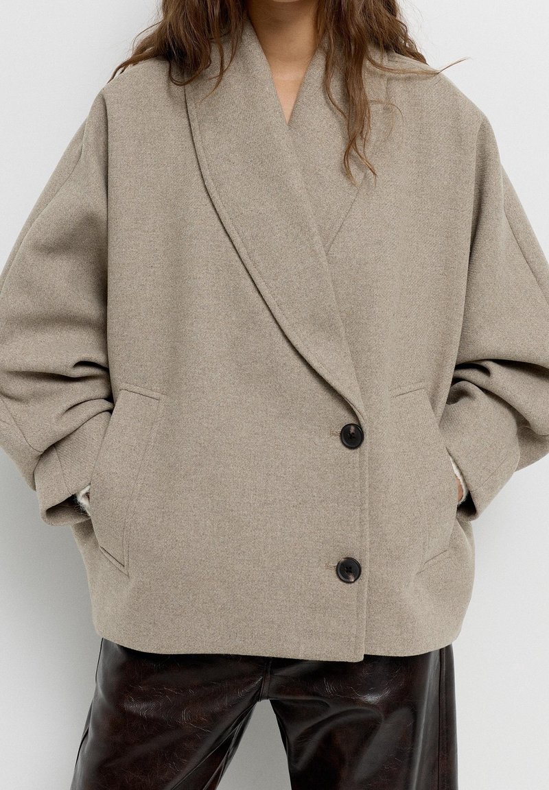 Femme portant un manteau en laine beige oversize avec deux boutons noirs sur un pantalon en cuir noir, les mains dans les poches du manteau.