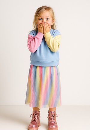 Pastellfarbener Kapuzenpullover mit rosa, blauen und gelben Ärmeln; plissierter Regenbogenkilt; glänzende rosa Kampfstiefel; Hände, die den Mund bedecken.