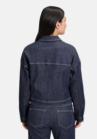 Tmavá džínová bunda s uvoľneným strihom, preloženým golierom a viditeľným šitím. Má zapínacie manžety a bočné vrecká.