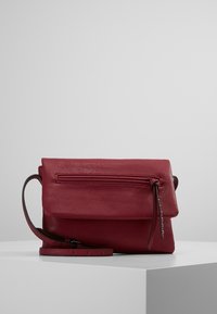 Červená kožená kabelka přes rameno s klopou, kapsou na zip a nastavitelným popruhem. Hladká textura s jednoduchými kovovými doplňky.