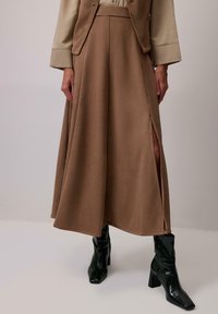 Barna midi szoknya sima textúrával és magas derékkal. Oldalsó hasítékkal rendelkezik, és fekete fényes bokacsizmával van párosítva.