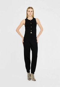 Schwarzes ärmelloses Oberteil mit durchgehender Knopfleiste, kombiniert mit schwarzen, zulaufenden Hosen. Das Modell trägt strukturierte Stiefeletten mit Schlangenprint.