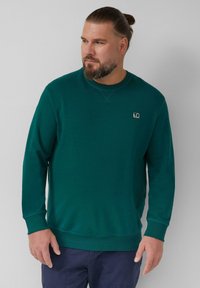 Turkusowa bluza z miękkiego materiału, posiadaj�ąca długie rękawy, okrągły dekolt oraz małą naszywkę z logo po lewej stronie klatki piersiowej. Gładka faktura.