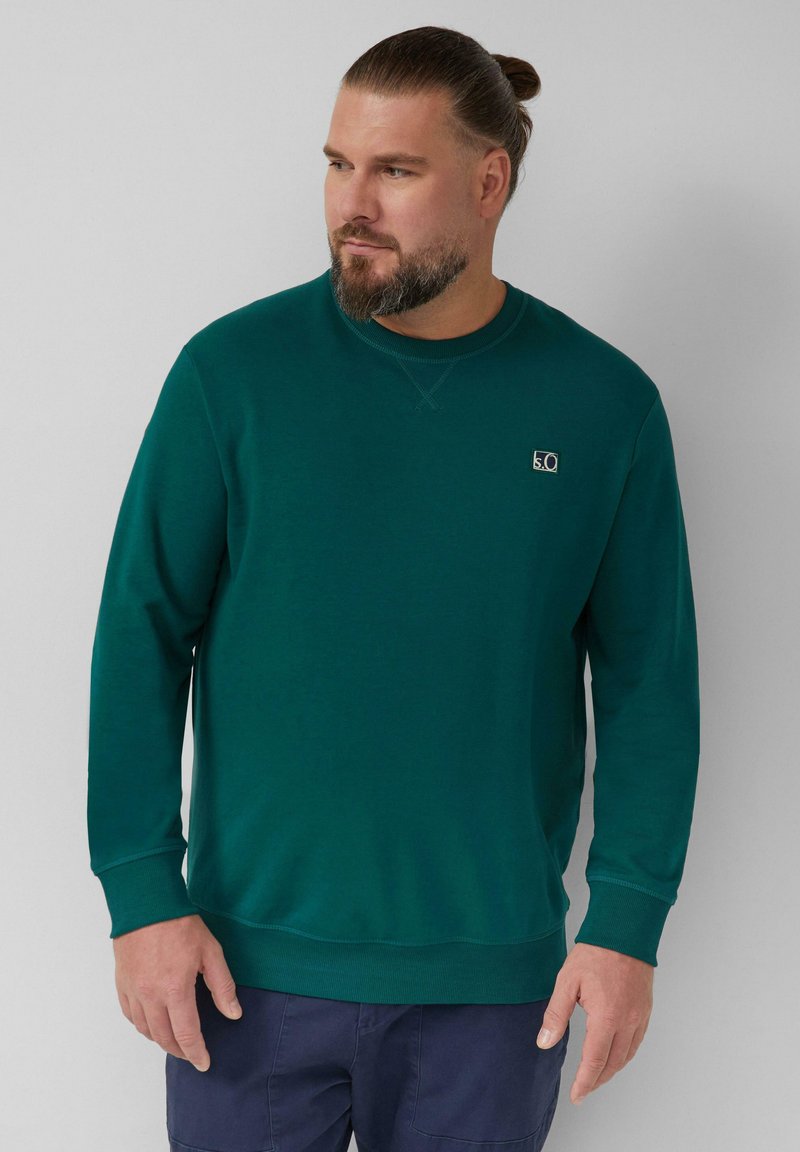 Turkusowa bluza z miękkiego materiału, posiadająca długie rękawy, okrągły dekolt oraz małą naszywkę z logo po lewej stronie klatki piersiowej. Gładka faktura.