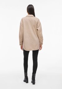 Beige oversized ingdzsekk, gallérral, gombos mandzsettákkal és kerek aljjal, fekete skinny farmernadrággal és bokacsizmával párosítva.