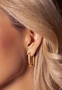 Gold hoop earrings with a rectangular design, smooth texture, and slight shine, worn on a model's ear with flowing blonde hair.