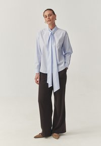 Light blue striped blouse with a tied neck detail, paired with loose-fitting dark brown pants. Soft, smooth fabric with long sleeves and pockets.