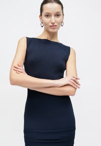 Γυναίκα με μαλλιά δεμένα πίσω φοράει αμάνικο μπλε φόρεμα με υφή στο τελείωμα, μεγάλα κρίκους σκουλαρίκια και σταυρώνει τα χέρια της με αυτοπεποίθηση απέναντι σε λευκό φόντο.