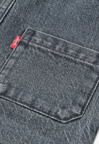Gros plan d’une poche arrière de jean en denim bleu avec des coutures visibles et une petite étiquette de marque en tissu rouge sur le côté.