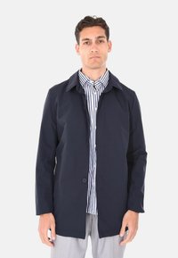 Giacca leggera di colore blu navy, con colletto, apertura con bottoni e maniche lunghe; indossata sopra una camicia a righe, realizzata in un tessuto liscio.