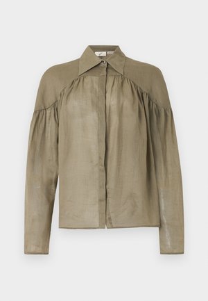 Femme portant un chemisier marron ample boutonné et une longue jupe blanche avec une fente devant, debout sur un fond blanc uni.