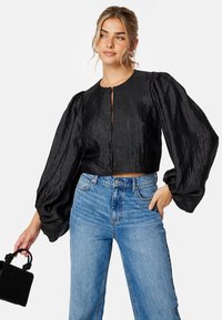 Zwarte cropped blouse met puffed mouwen, voorzien van een ronde halslijn en sluiting aan de voorkant. Gecombineerd met lichtblauwe wijde jeans en een gestructureerde zwarte tas.