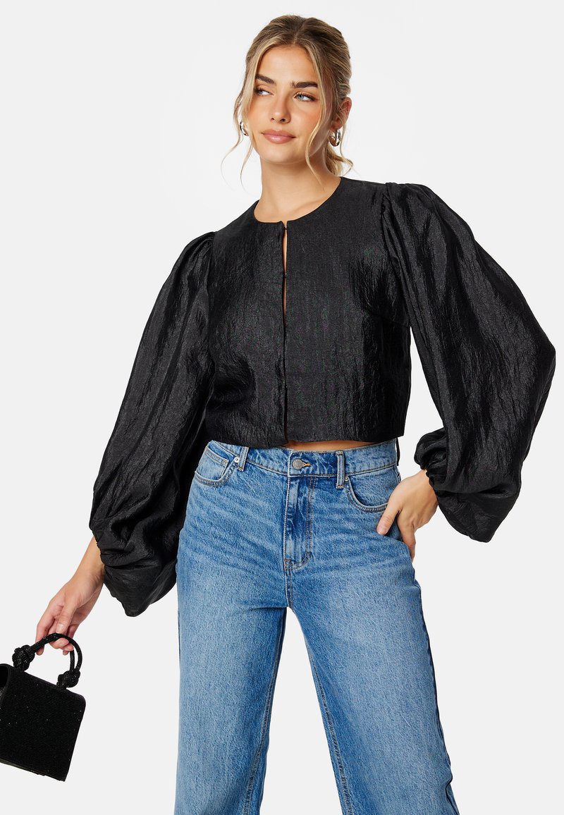 Zwarte cropped blouse met puffed mouwen, voorzien van een ronde halslijn en sluiting aan de voorkant. Gecombineerd met lichtblauwe wijde jeans en een gestructureerde zwarte tas.