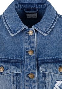 Chaqueta de denim en tono azul claro, con cuello clásico, botones dorados y dos bolsillos en el pecho. Tejido texturizado con costuras contrastantes.
