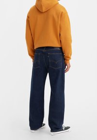 Person som bär mörkblå vida jeans, en senapsgul hoodie och svartvita sneakers, stående mot en vit bakgrund.