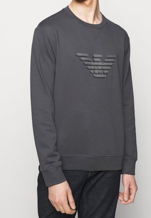 Homme portant un sweat-shirt à manches longues gris foncé avec un logo aigle à rayures horizontales texturées sur la poitrine, assorti à un jean foncé.