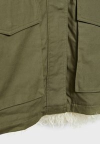 Olivgrüner Oberstoff mit großen Klappen Taschen und weichem weißen Lammfellfutter am Saum, das eine glatte Textur und saubere Nähte zeigt.