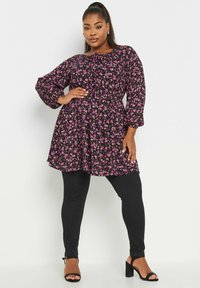 Blommig tunika i svart med rosa blommor, rund halsringning, långa volangärmar och en A-linjeformad silhuett, matchad med svarta leggings och klacksandaler.