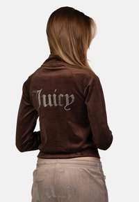 Giacca in velluto marrone con "Juicy" in strass sul retro. Abbinata a pantaloni beige con un dettaglio simile in strass. Tessuto morbido.