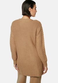 Long cardigan en tricot dans un brun clair. Présente un design texturé avec un motif en torsades aux manches et une coupe décontractée.