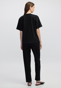 Černé tričko s krátkým rukávem a kulatým výstřihem, v kombinaci s černými slim-fit kalhotami a plochými černými botami, viděné zezadu.