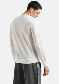 Maglione bianco a maniche lunghe con design a maglia texture, collo rotondo e orlo a costine. Indossato con pantaloni grigio scuro con strisce bianche.
