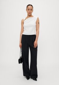 Witte top met één schouder, vervaardigd uit een gladde stof, gecombineerd met zwarte pantalon met wijde pijpen. Het model houdt een kleine zwarte handtas vast, met geometrische plooien.