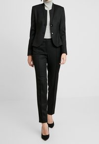 Zwarte op maat gemaakte blazer met een cropped jack, twee knopen en slim-fit broek. Gdragend met een lichtgrijze coltrui en zwarte hakken.
