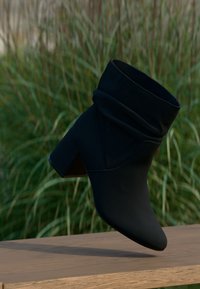 Bottine noire à bout pointu, en matière douce et texturée, tige souple, et talon carré, présentée devant un fond de verdure floue.