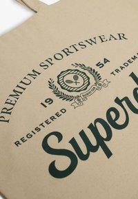 Beżowa torba tote z płótna z ciemnozielonym nadrukiem. Zawiera branding "Premium Sportswear" oraz "Superdry", a także dekoracyjne elementy i wzory.