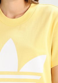 Yellow cotton t-shirt with a crew neck, featuring a large white graphic of three leaves and horizontal stripes across the chest.