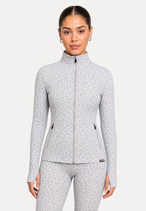 Mujer con una chaqueta gris claro con cremallera y leggings a juego con un sutil estampado de leopardo, de pie frente a un fondo liso.