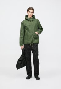 Χακί πράσινο μπουφάν με κουκούλα και κορδόνι, μεγάλο μπροστινό τσέπη, συνδυασμένο με υφασμένα μαύρα παντελόνια. Το μοντέλο κρατά μια μαύρη καπιτονέ τσάντα.