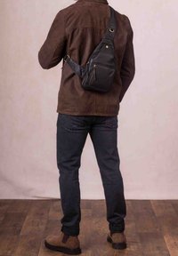Black leather sling bag with zippered pockets, adjustable strap, and gold accents, worn over a brown suede jacket and dark jeans.