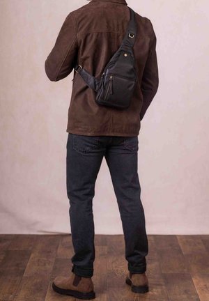 Black leather sling bag with zippered pockets, adjustable strap, and gold accents, worn over a brown suede jacket and dark jeans.