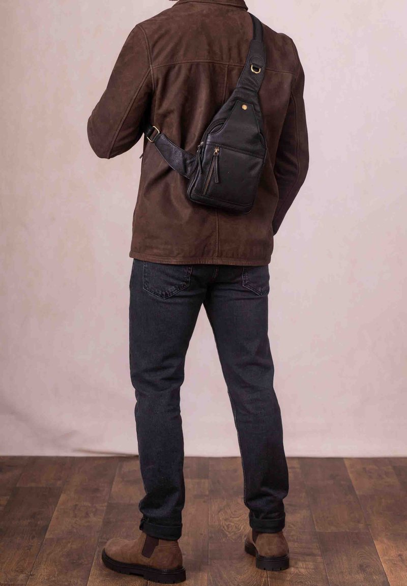 Black leather sling bag with zippered pockets, adjustable strap, and gold accents, worn over a brown suede jacket and dark jeans.