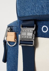 Sac à dos en denim avec une sangle navy et une boucle noire gravée "TOMMY JEANS". Comprend une étiquette en cuir tan et des fermetures éclair en argent.