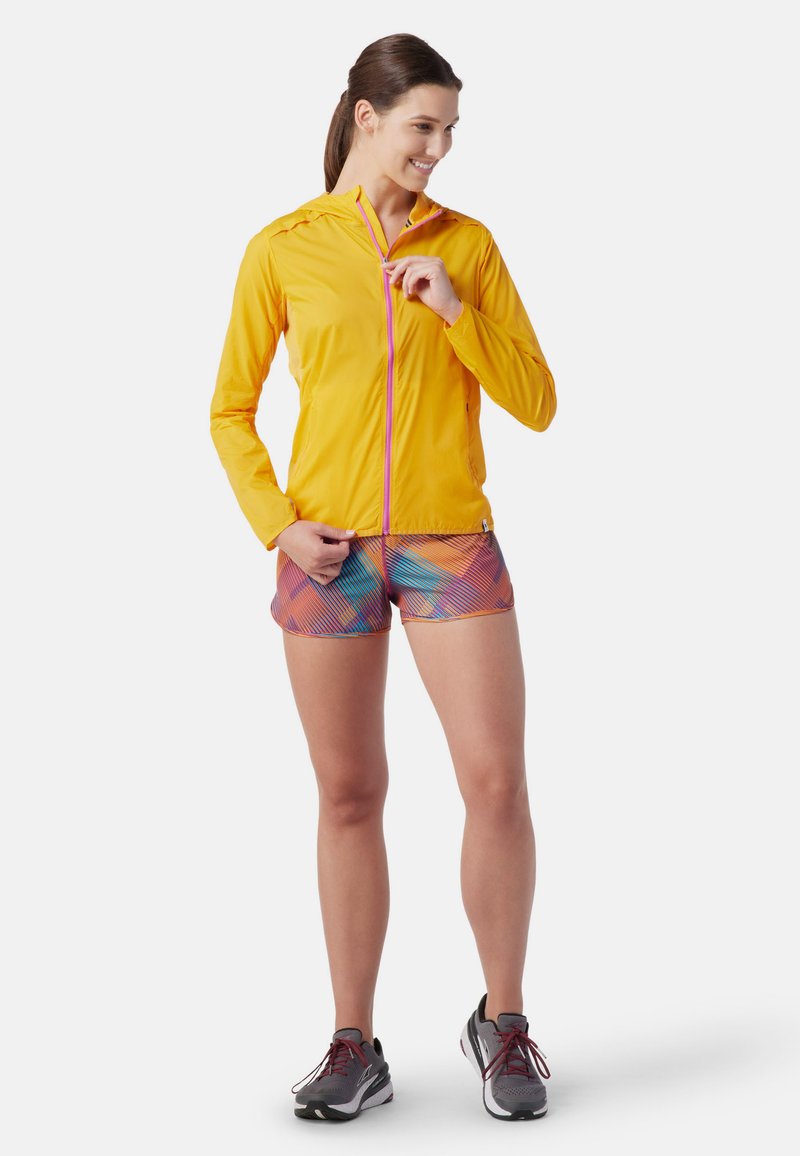 Lätt gul jacka med rosa dragkedja, tillsammans med flerfärgat mönstrade shorts. Modellen har på sig grå sportskor och står avslappnat.