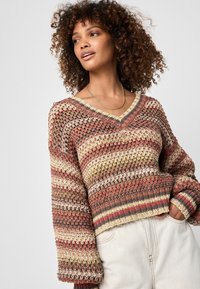 Femeie îmbrăcată într-un pulover tricotat în culori multiple, cu decolteu în V și mâneci lungi, purtând pantaloni deschiși la culoare, cu talie înaltă, cu mâna în buzunar.